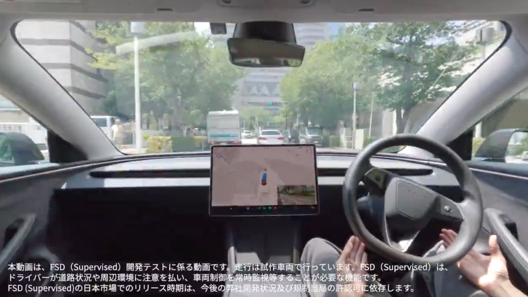 テスラFSD自動運転テスト走行の車内映像テスラ車の運転席から撮影された映像で、ドライバーの両手がハンドルから離されており、自動運転中であることを示しています。右側にはテスラ特有の大型タッチスクリーンディスプレイが設置されており、画面には車両周辺の状況を示すグラフィカルな表示が映っています。フロントガラス越しには日本の都市部の街並みが見え、街路樹が並ぶ道路、他の車両、建物などが確認できます。車内のインテリアはテスラらしいミニマルなデザインで、黒を基調とした内装となっています。画面下部には日本語で以下のテロップが表示されています：「本動画は、FSD（Supervised）開発テストに係る動画です。走行は試作車両で行っています。FSD（Supervised）は、ドライバーが適切な注意力を払い、周辺を常時監視し、必要な場合にはいつでも運転を引き継ぐ必要があります。FSD（Supervised）の日本市場でのリリース時期は、今後の許可申請状況及び規制当局の許可可に依存します。」この映像は、テスラが日本でのFSD技術の実証実験を行っていることを示すものです。